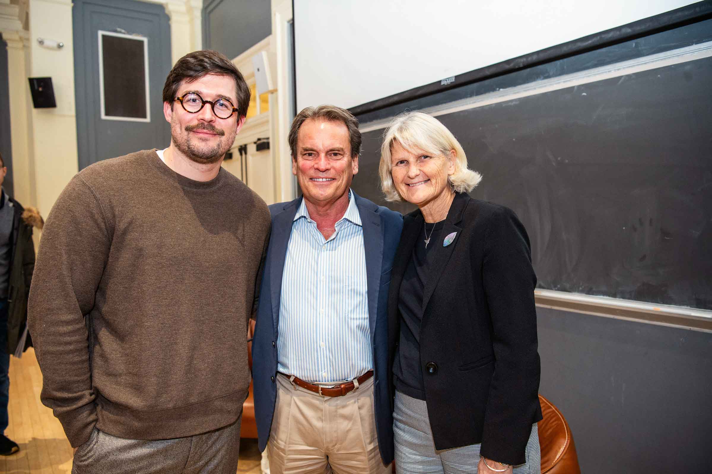 Three smiling people arm in arm standing in front of a chalkboard.