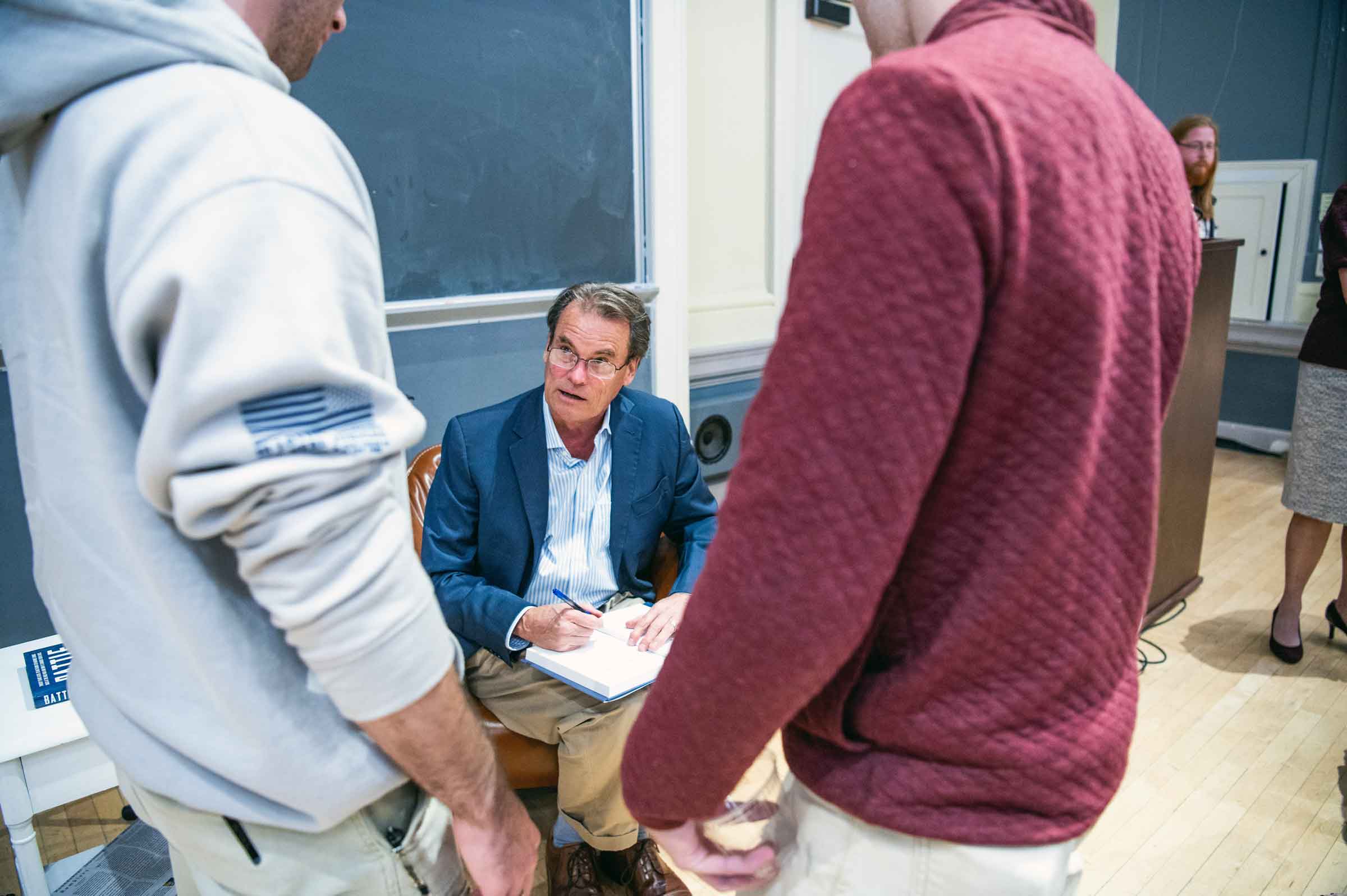 A person sitting in a chair signing a book and talking to two standing people.
