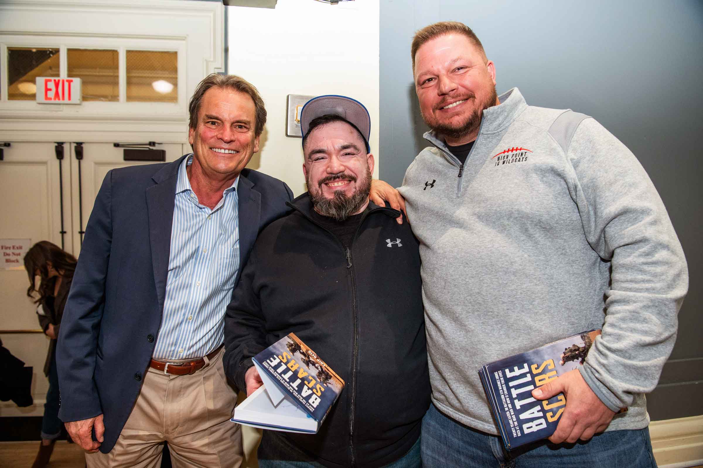 Three smiling people standing arm in arm. Two are holding books with a title that read: Battle Scars.