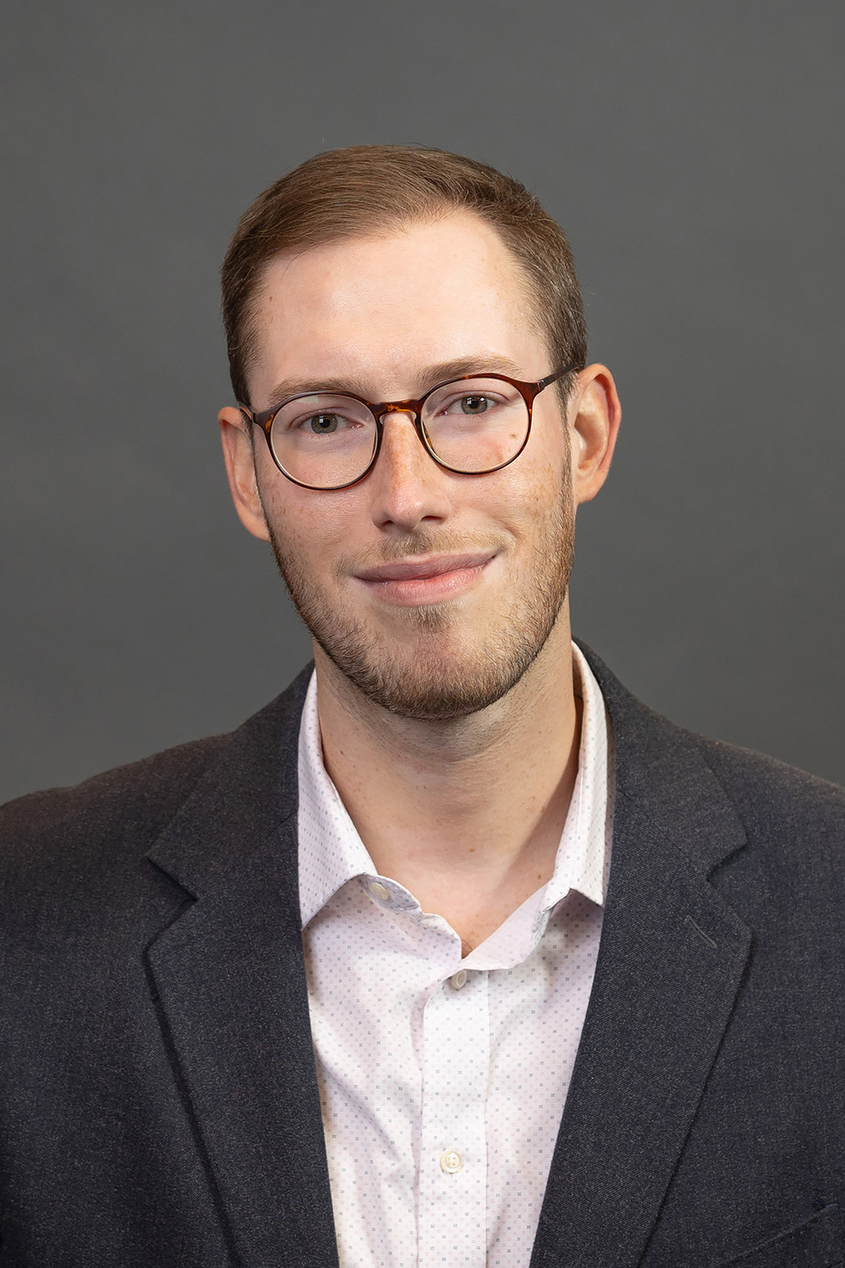 Person with brown short brown hair, tortoise shell glasses, white shirt with black blazer, smiling for a portrait. 