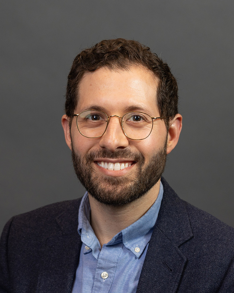 Headshot of person with dark curly short hair, glasses, and beard wearing a dark suit jack and light blue collared shirt.