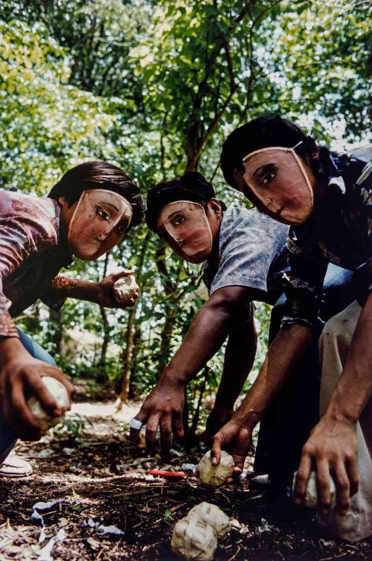 Three people wearing masks of human faces crouch on the floor of what looks like a forest, holding objects that they are preparing to throw.