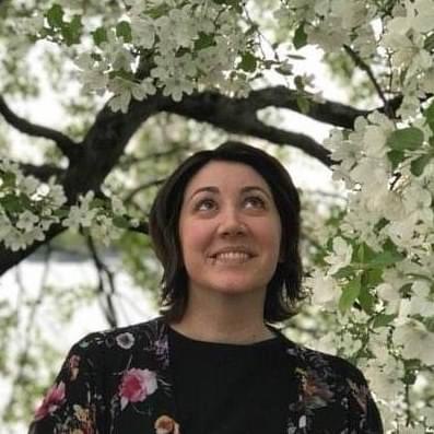 A person with long dark brown hair stands under a tree with white flowers, smiling off-camera.