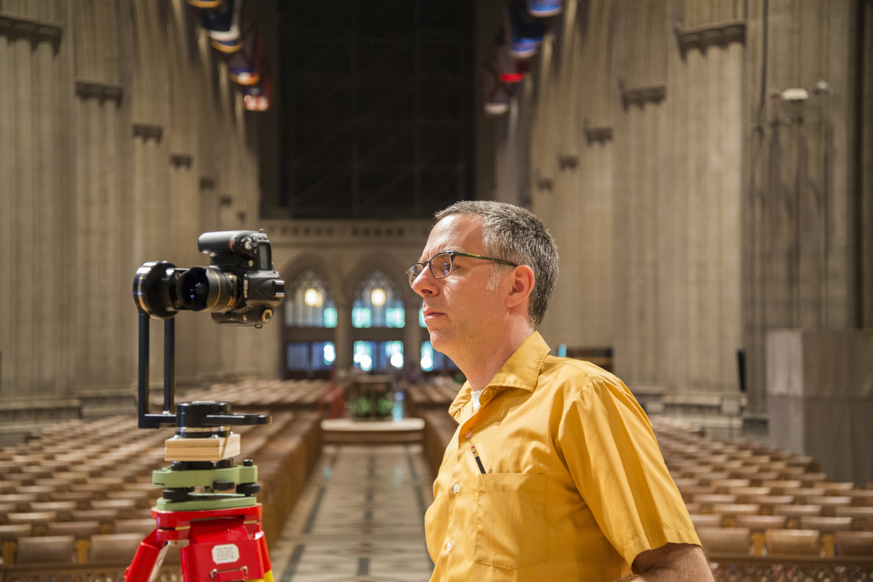In a nave, a man with short dark hair, wearing glasses and a bright yellow short sleeve shirt, is looking intently at a camera view screen with pews in the background.