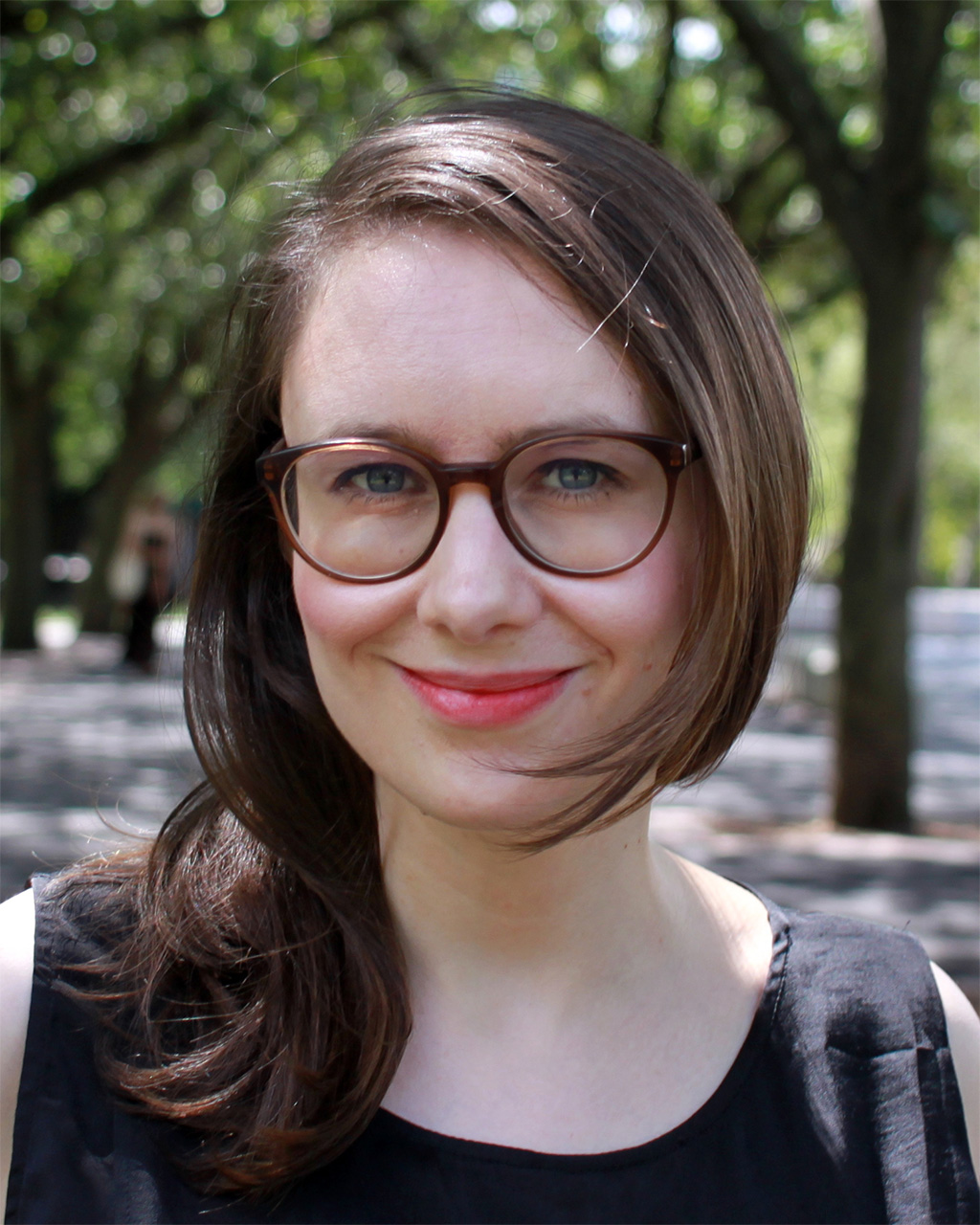 A woman with medium-length brown hair, wearing glasses and a black sleeveless shirt, smiling at the camera. The background shows a pathway lined in trees with green leaves.