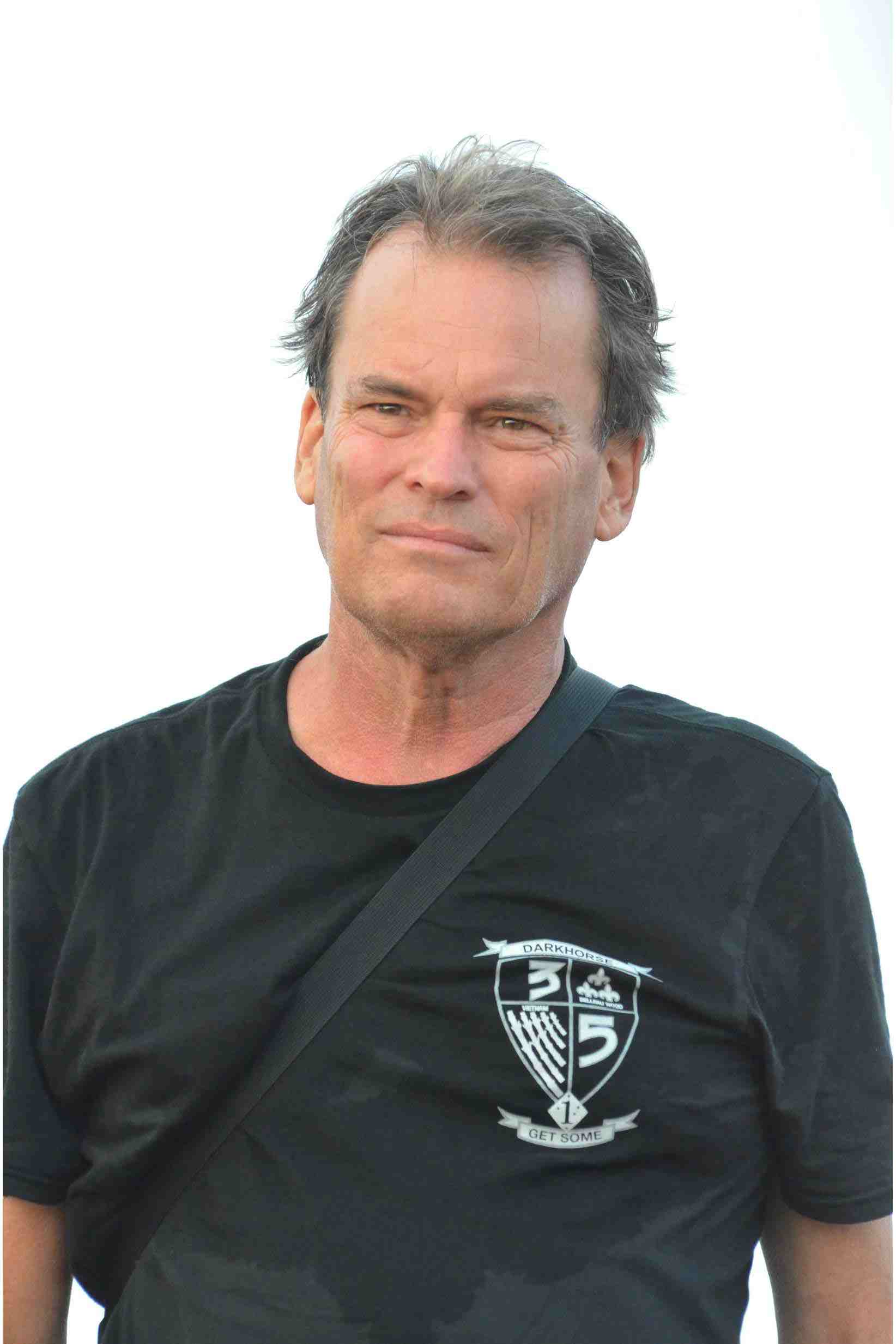 A photograph of a person with mid-length gray hair, a somewhat grim expression, and a black T-shirt with a small shield design in the top left.