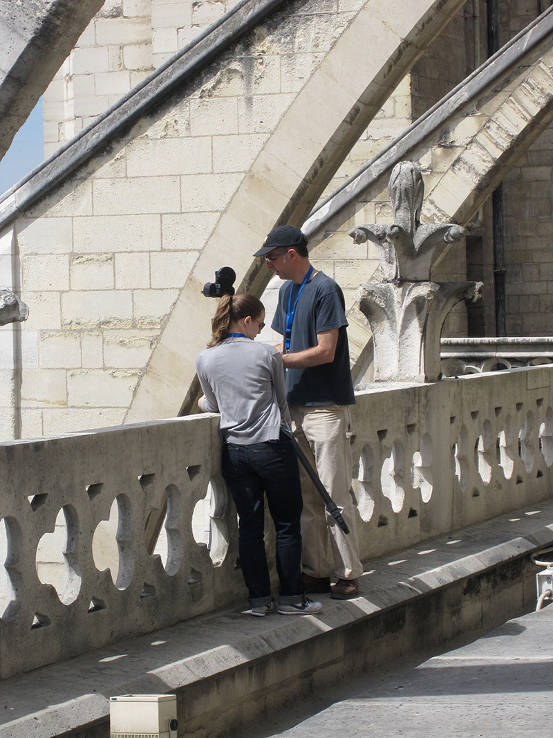 Two people with camera equipment setting up along a waist-high stone wall under stone arches.