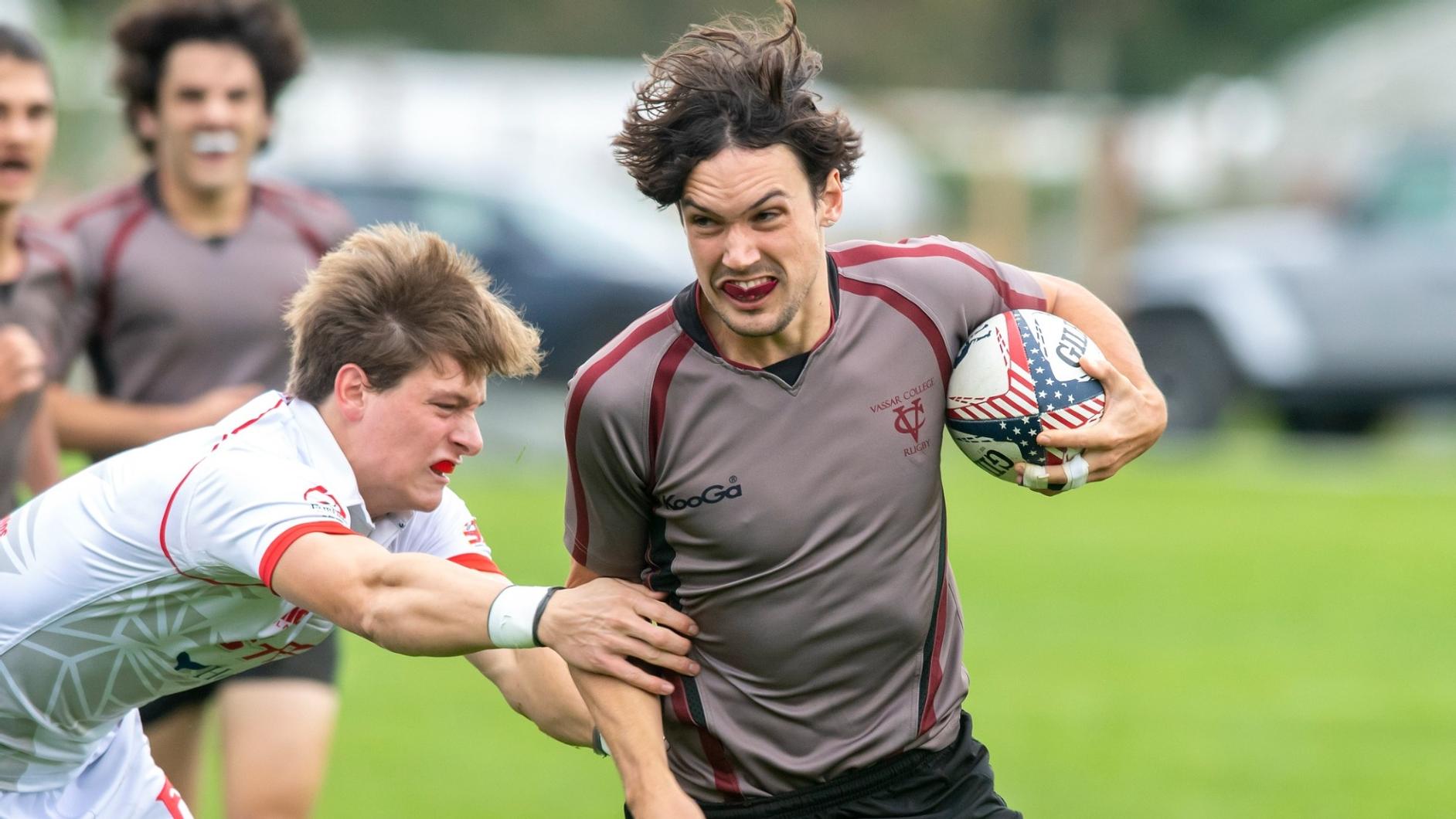 One rugby player holding the ball sprints past another player who is diving to tackle that player
