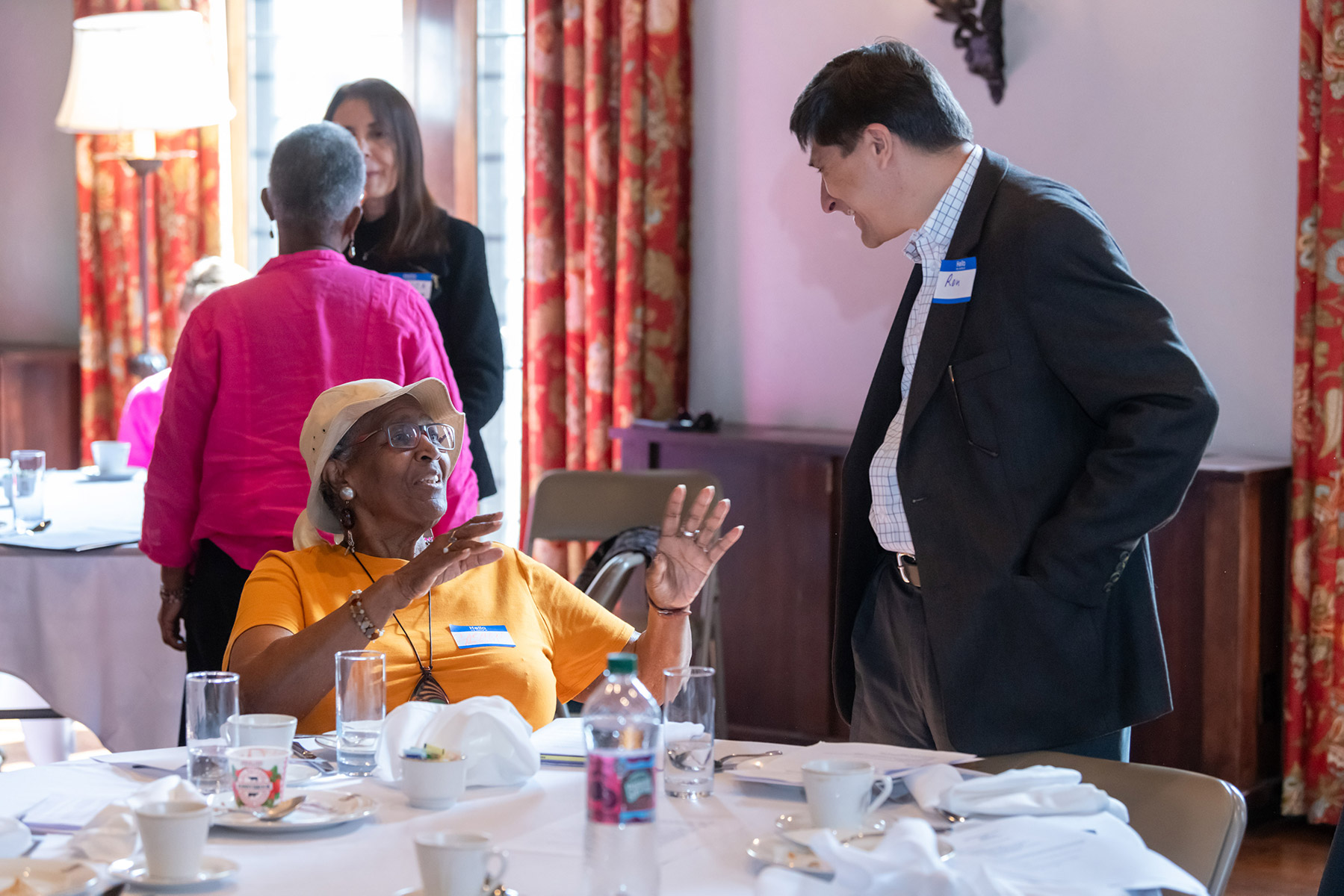 A person sitting at a table with plates and glasses on it is speaking to someone standing next to them, at the right of the photo. The seated person has glasses, a white hat, and an orange shirt. The standing person has short dark hair and a dark suit with a nametag that says “Ron”. Two people are standing in the background talking.