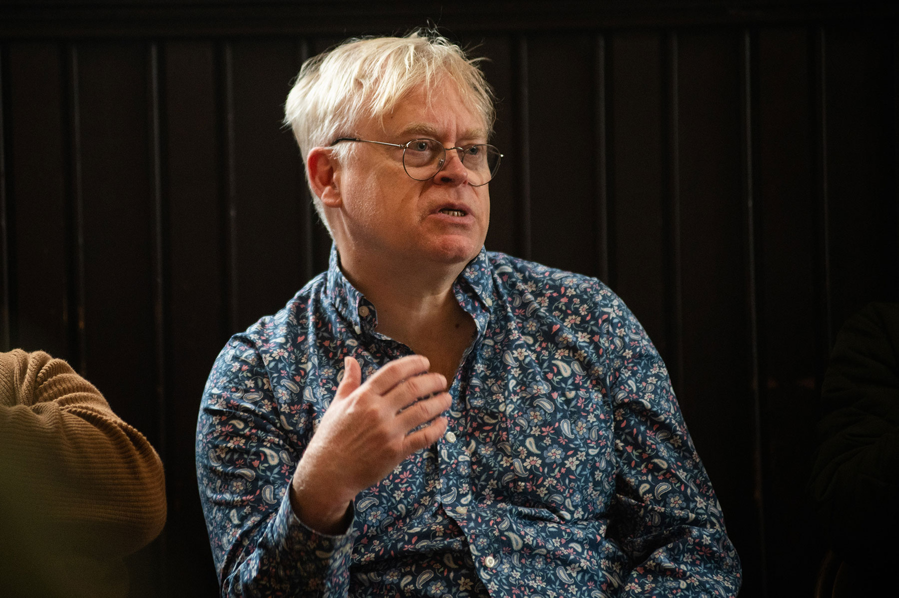 Person with white blonde hair, glasses, and a blue shirt sitting on a stage speaking.