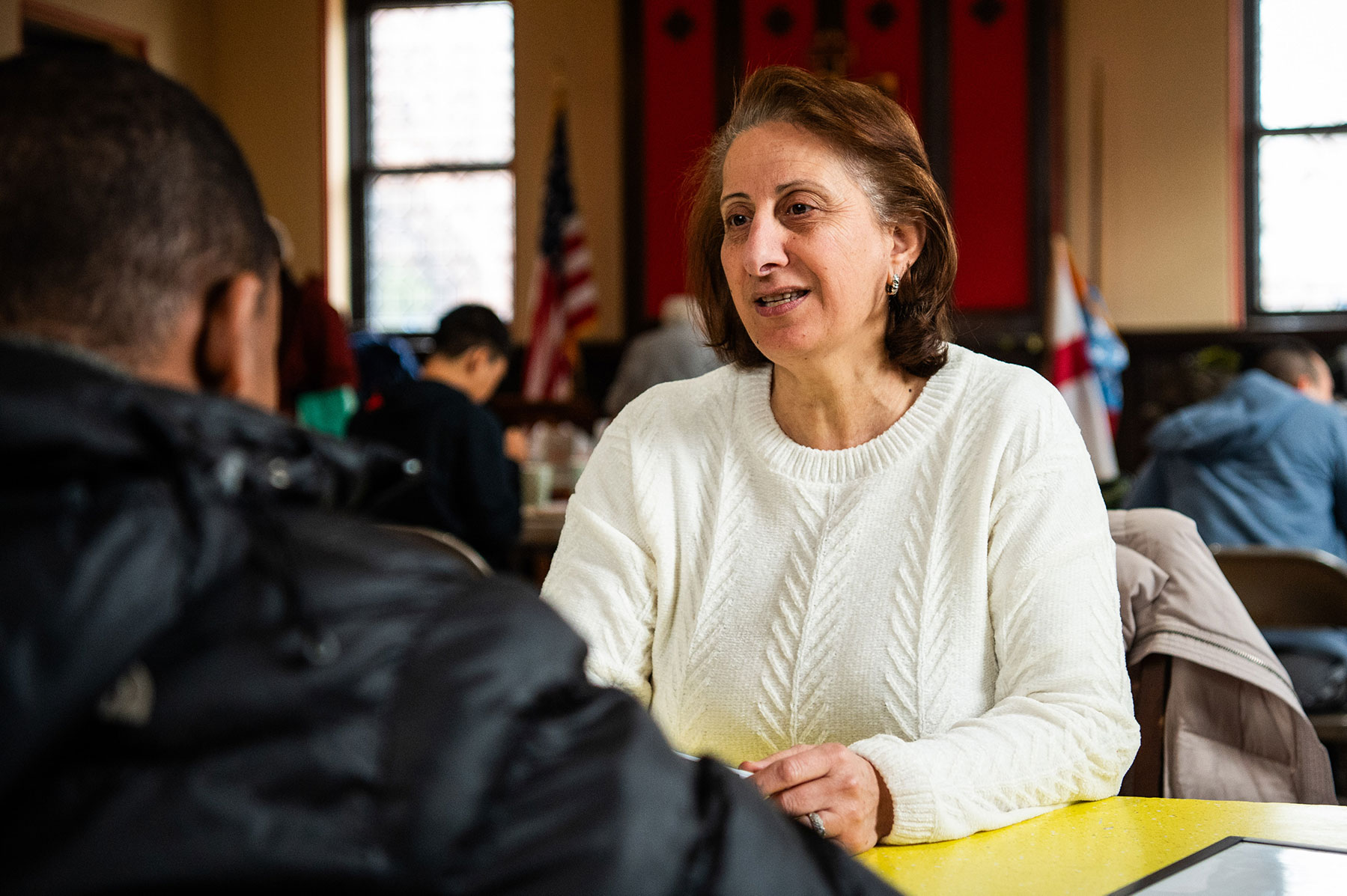 Person with a hat sitting and talking to another person across from them at a white table.