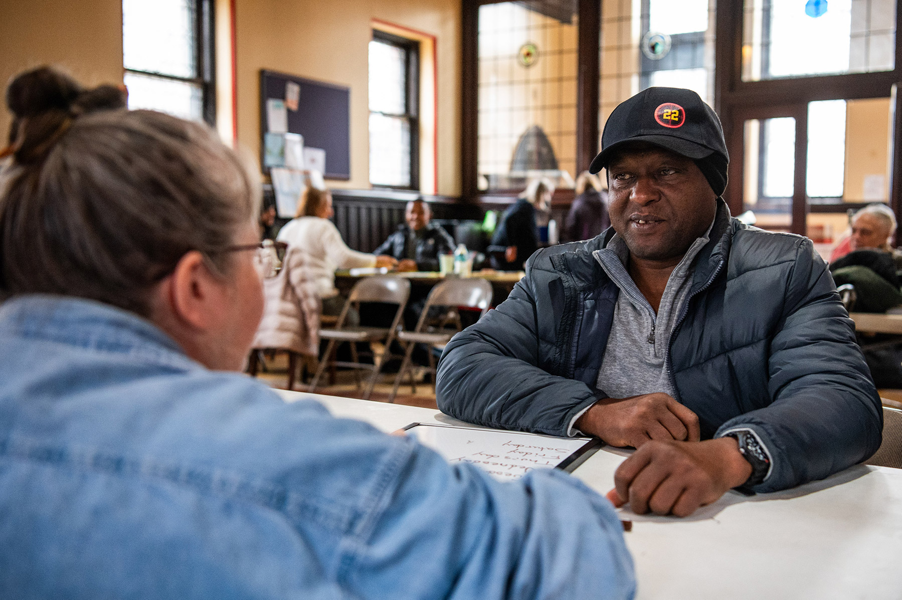 Person with a hat sitting and talking to another person across from them at a white table.