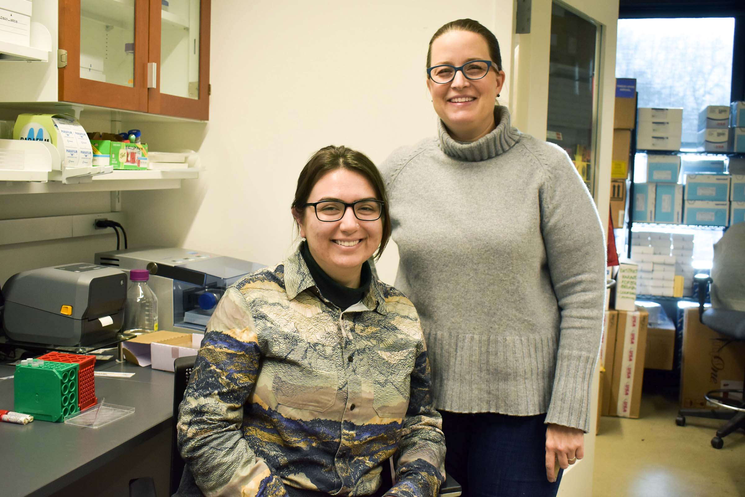 Morgan Stephens ’23 (left), sitting and wearing a printed collared shirt, and Visiting Scholar Elizabeth Thiele, standing and wearing a gray turtle neck sweater, in a research lab.
