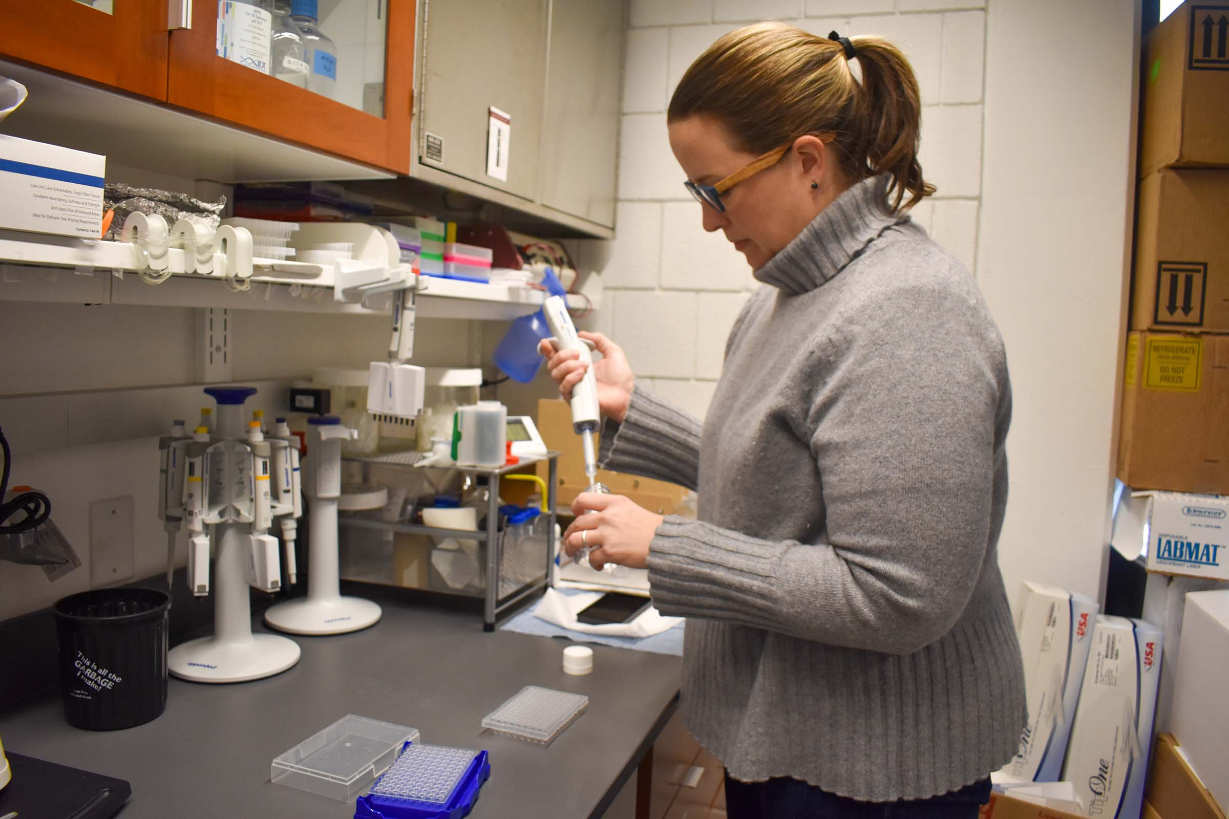 Elizabeth Thiele wearing a gray turtleneck sweater, standing in a lab and holding a large syringe in a glass vial.