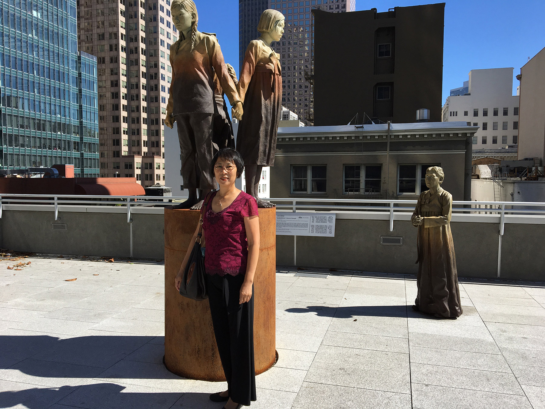 Person posing on a city rooftop in front of three bronze statues of people in different poses.