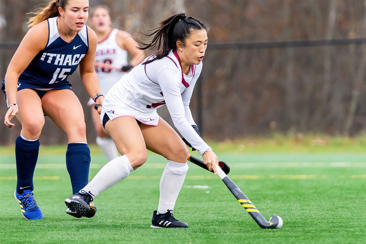 Juliana Ishimine hits the ball with her stick while being guarded from behind by an opponent.