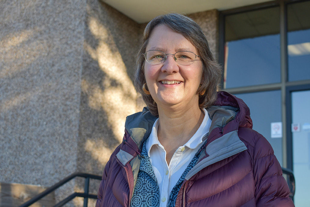 A person with salt-and-pepper hair wearing glasses and a purple winter jacket standing outdoors, smiling.. 