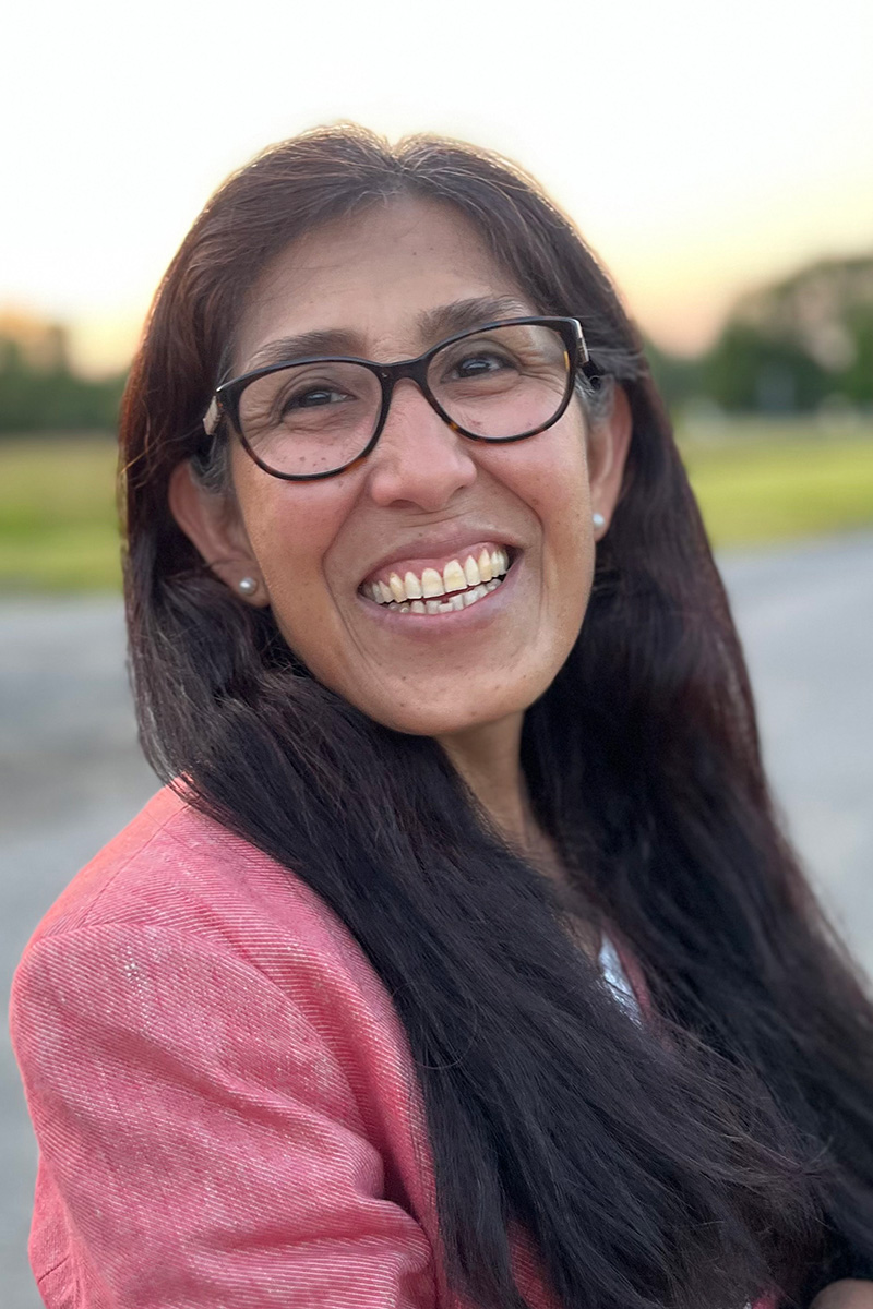 A person with long dark hair, brown skin, wearing glasses broadly smiling.