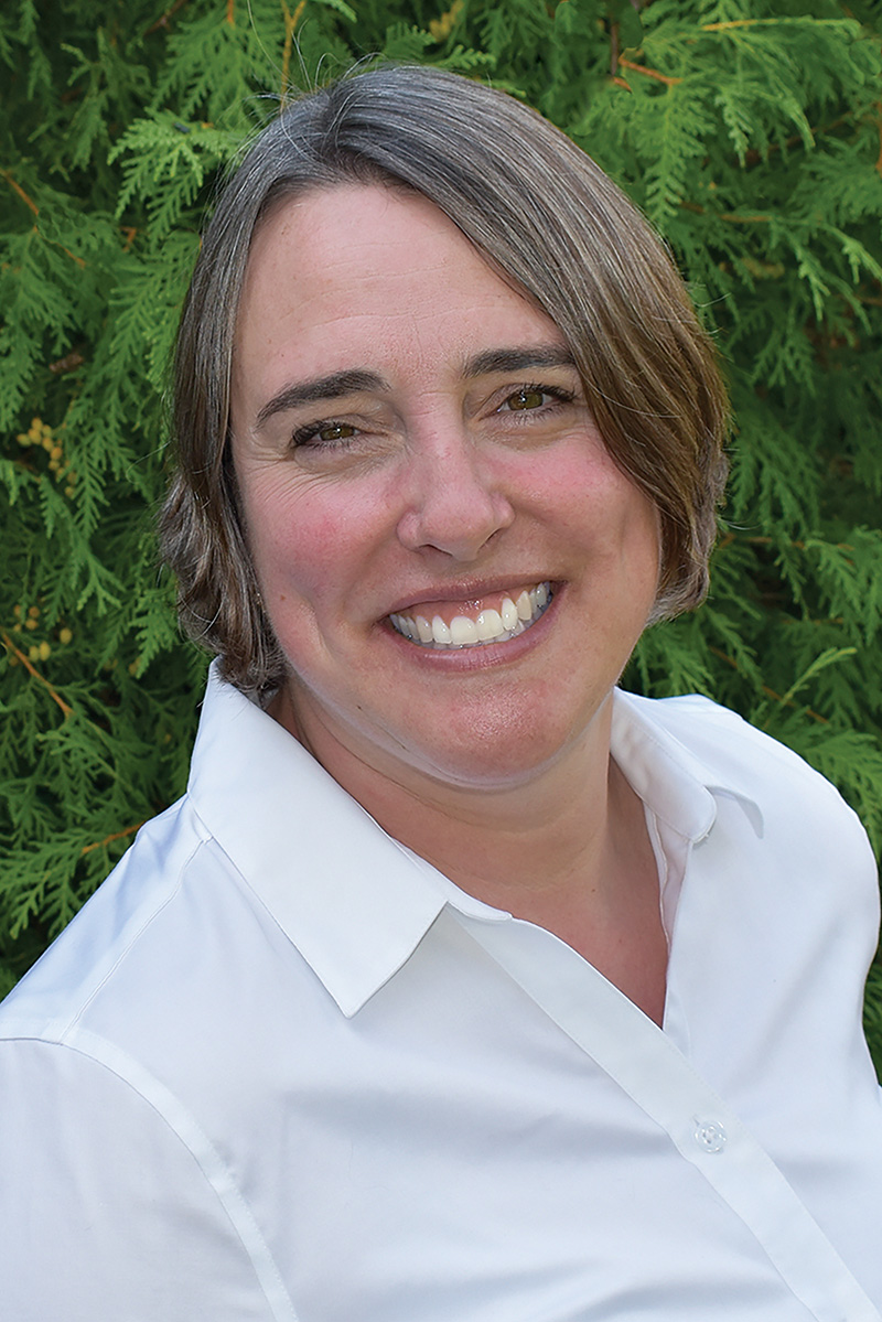 A person with medium length dark blonde straight hair wearing a white collared shirt facing the camera smiling.