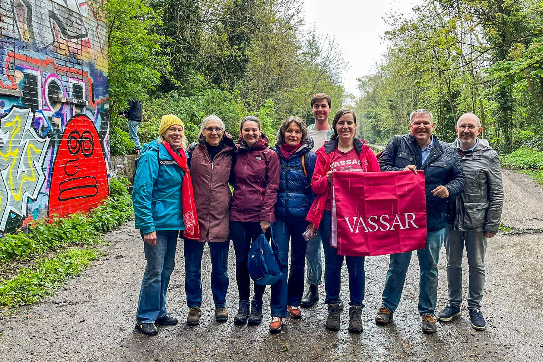 A group of people standing together on a gravel road in a wooded area holding a Vassar banner