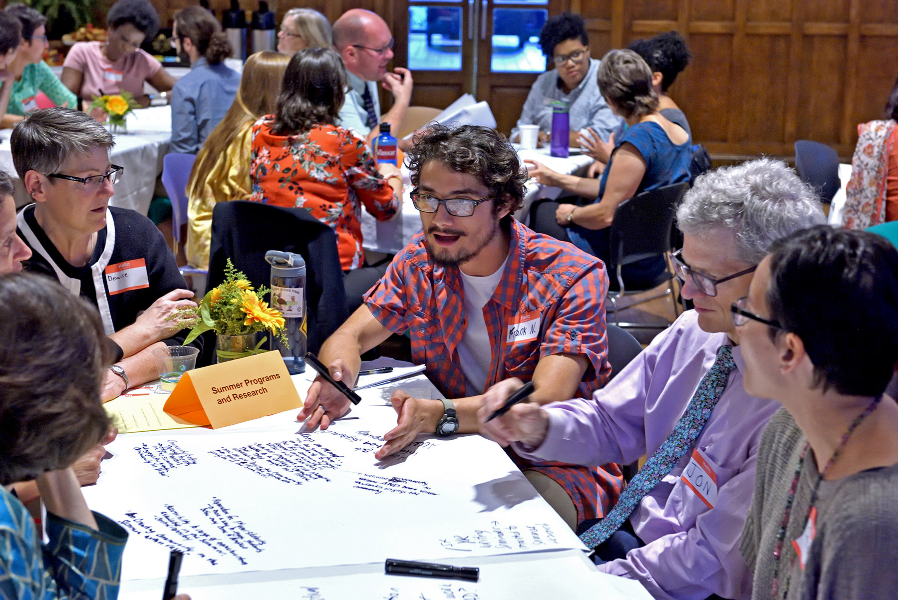 Group of people sitting at a table talking with notes on the table. The background room is filled with other round tables with people seated talking.