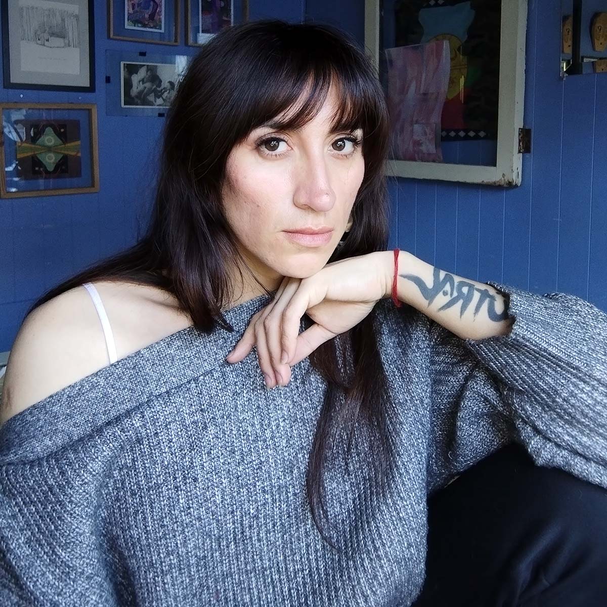 Kütral Vargas Huaiquimilla wearing a blue and white knit sweater sitting in front of blue walls with various artwork.