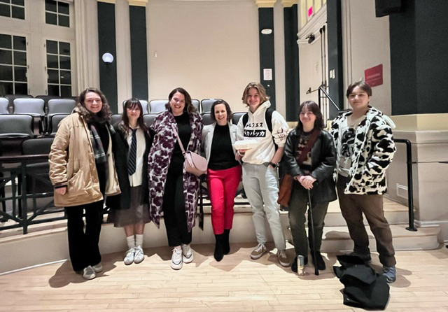 Alum Lynne Marie Rosenberg standing with six students in front of black theater seating.