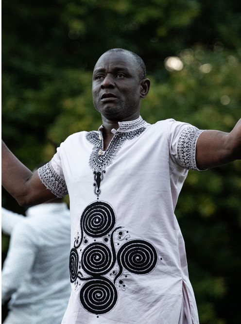 A person with short black hair and a white shirt with geometric spiral patterns on it stands outside, arms apart. They look like they are either teaching or performing.