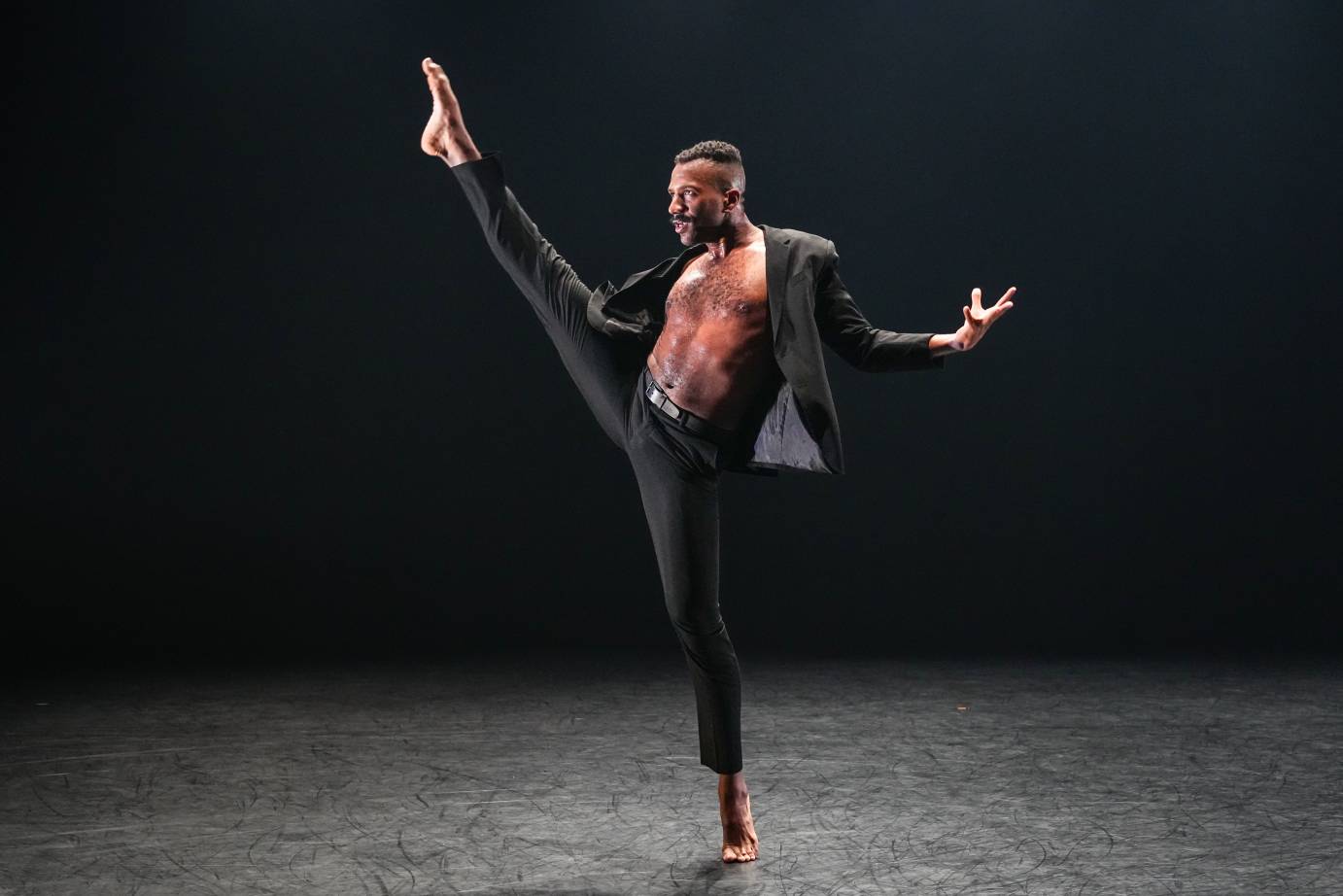 A person wearing a black suit jacket and dress pants dances on a dark stage, one foot raised high in the air.