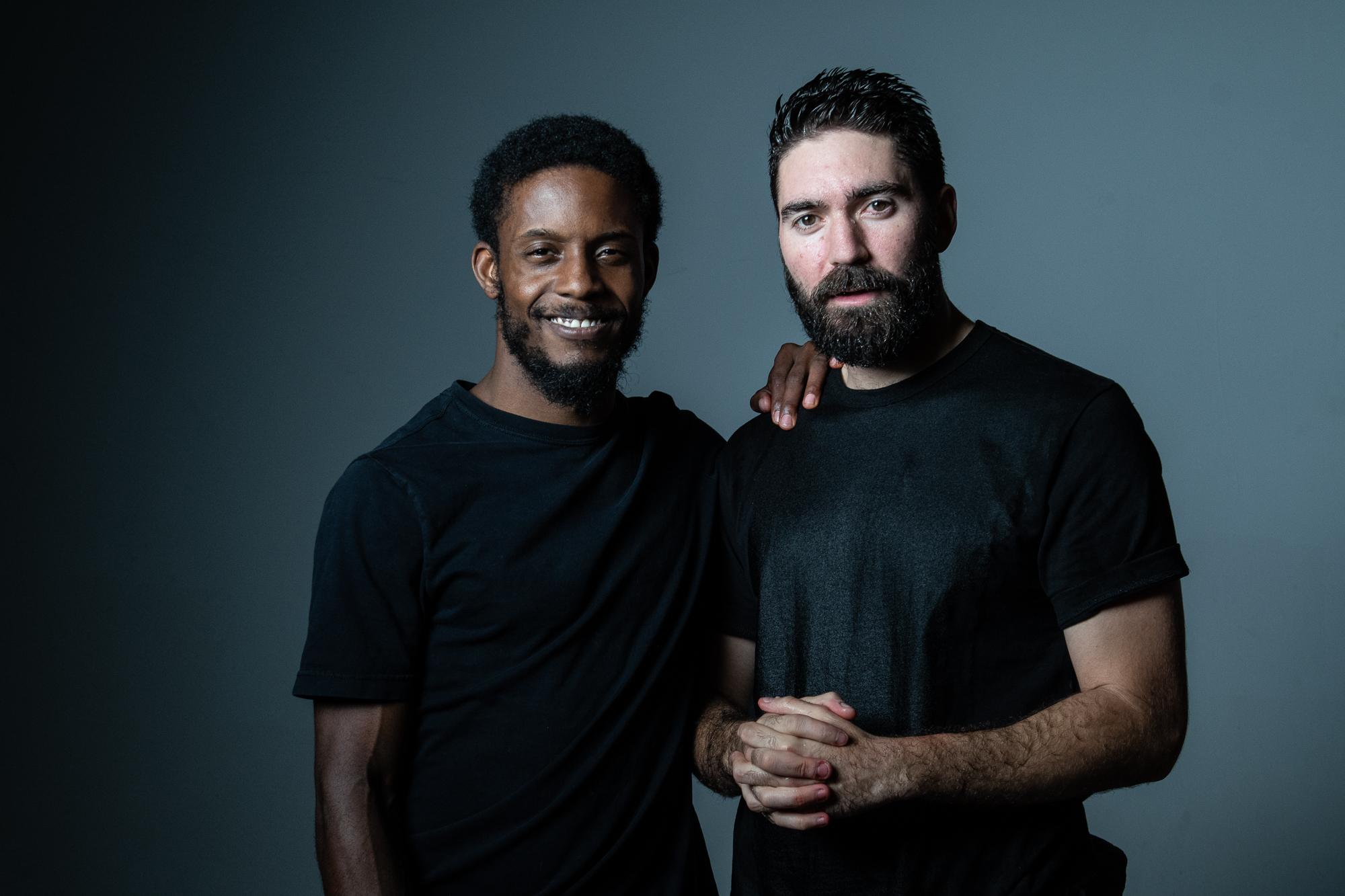 Two people wearing dark T-shirts stand together in a darkened room, smiling at the viewer.