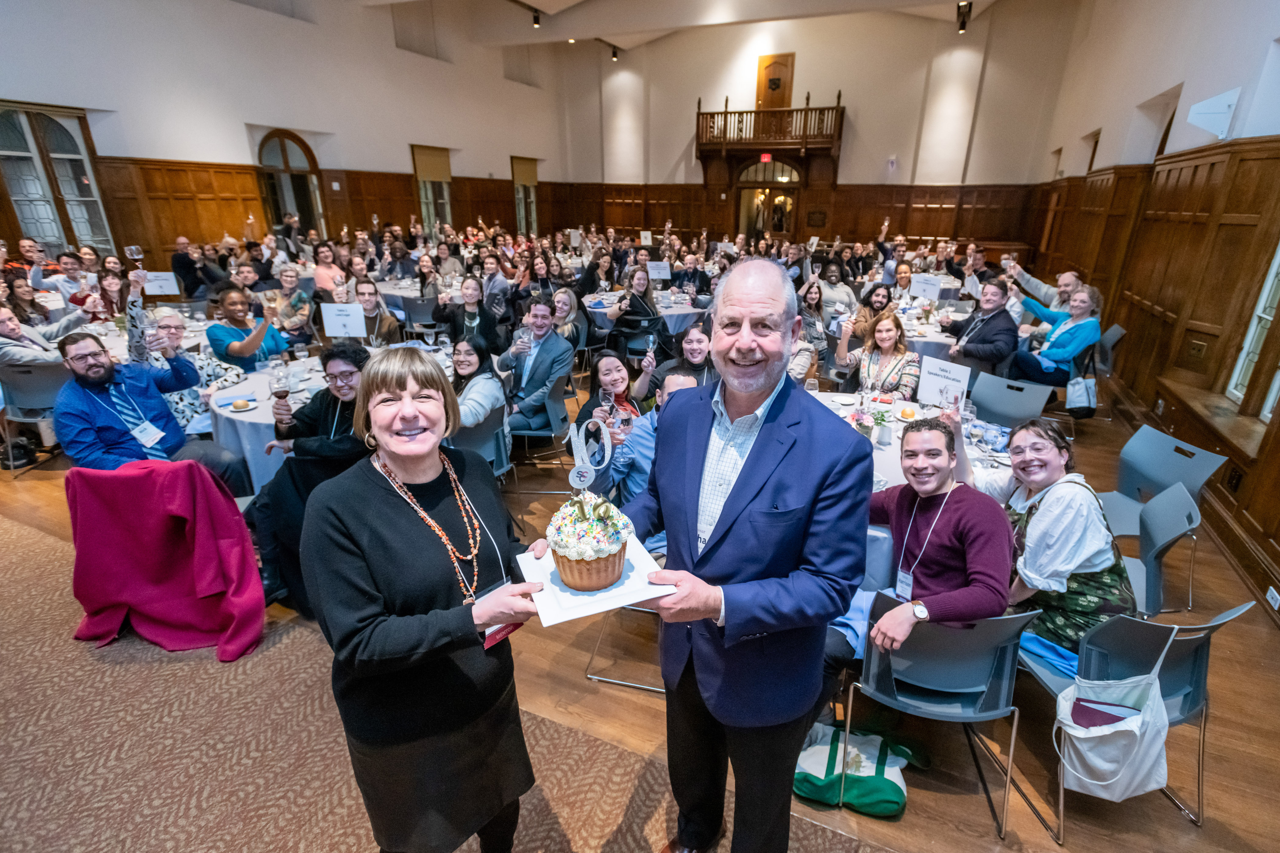 Two people facing the camera presenting a plate with a large frosted cupcake. They are standing in front of a large room full of people seated at round tables, all people facing the camera.