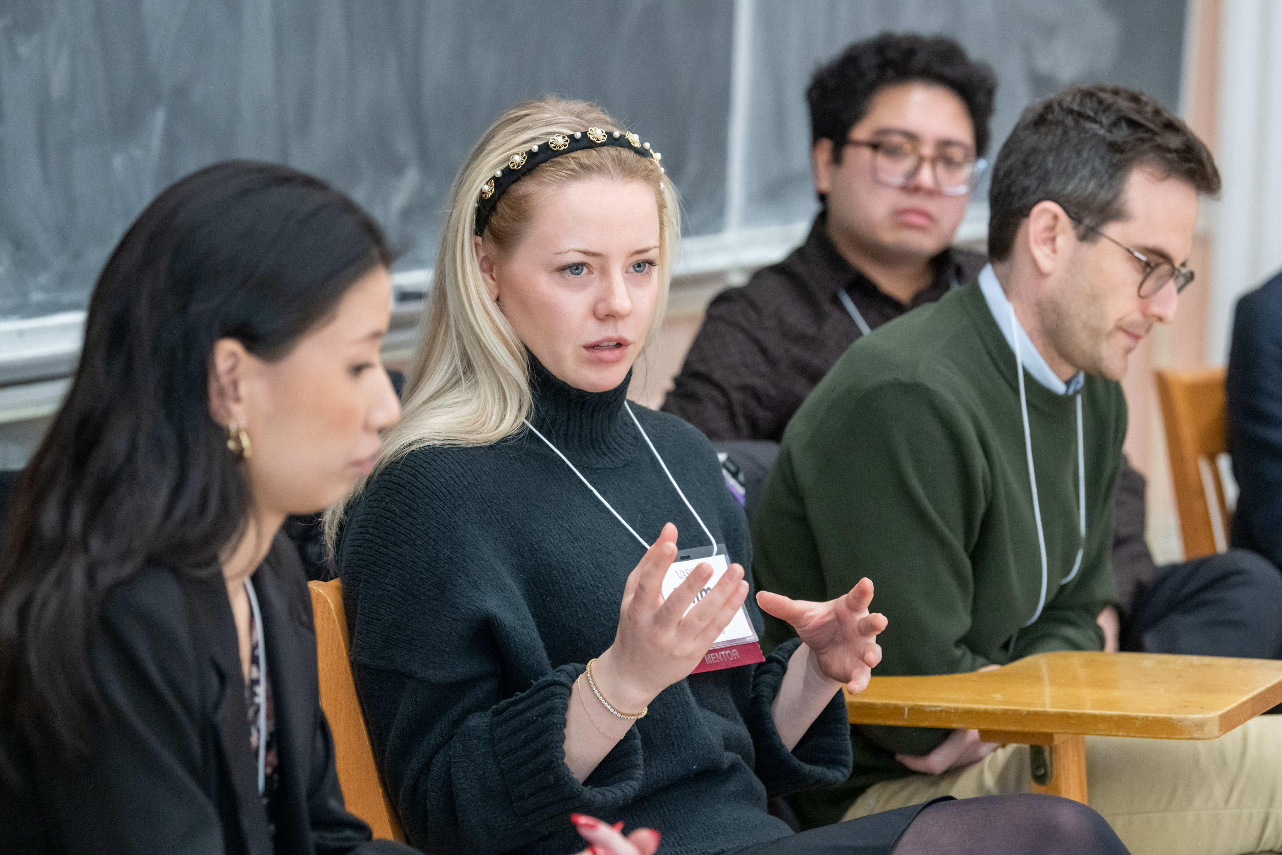 Four people sitting in front of a chalkboard, with one person in the center speaking, motioning with their hands.