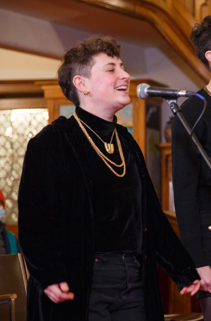 A person with short hair wearing a black long-sleeve shirt and gold necklace singing with a standing microphone.