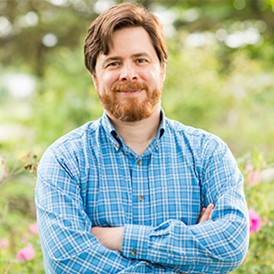 portrait of Peter Henne standing outdoors with his arms crossed, with with tall grass and flowers in the background..