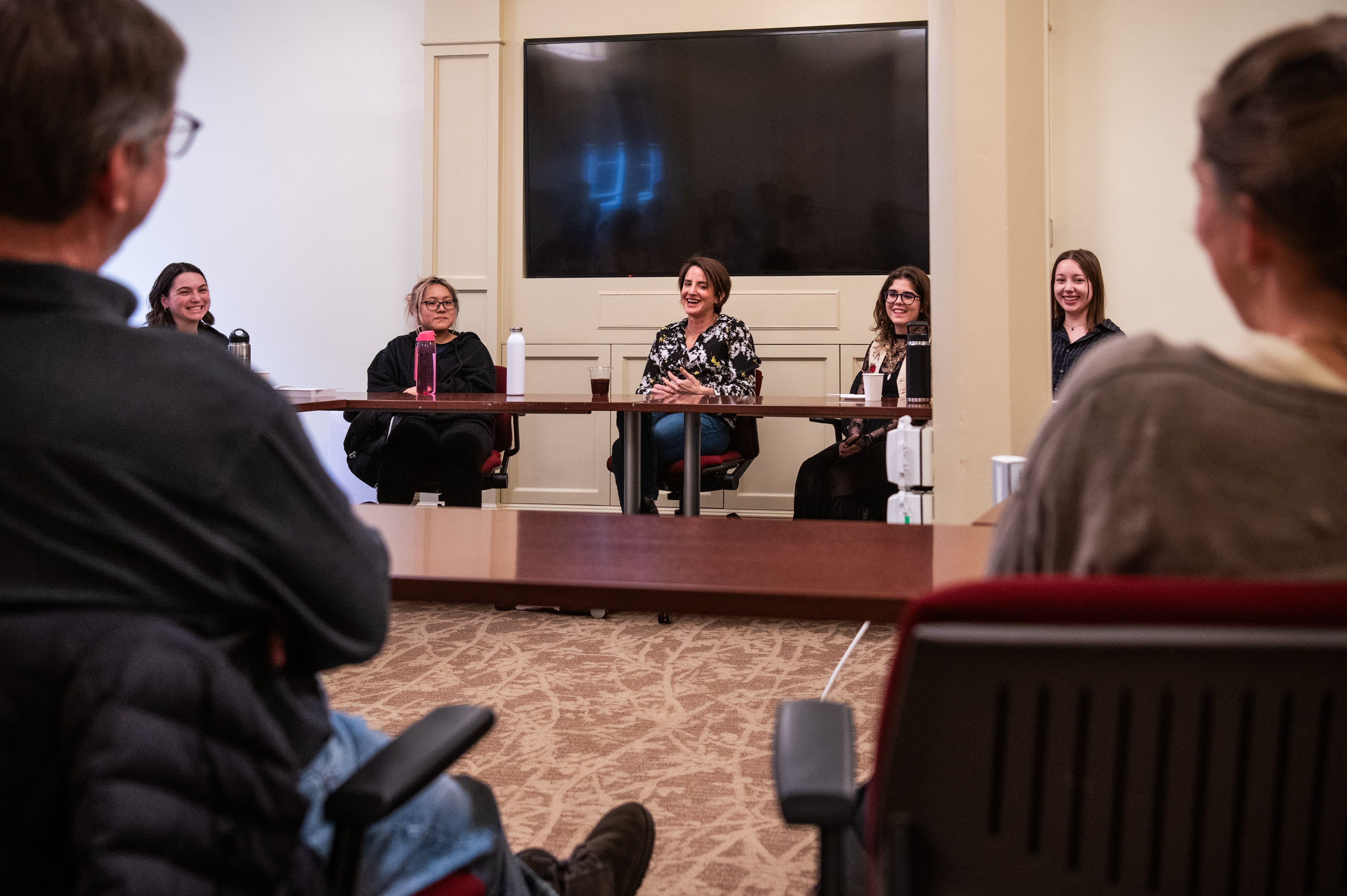 Two seated people with backs to camera watching and listening to a panel discussion of seated people behind a table.