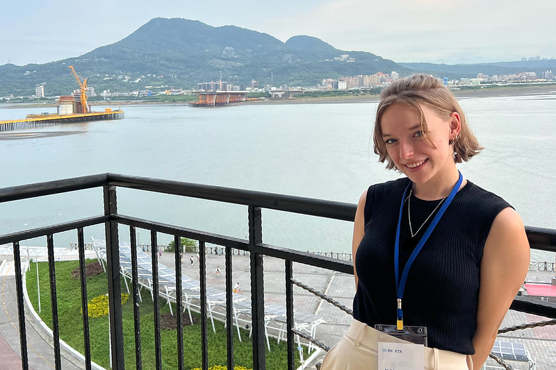 Person standing on a balcony with metal railing with and overlook of a river and small mountain backdrop.