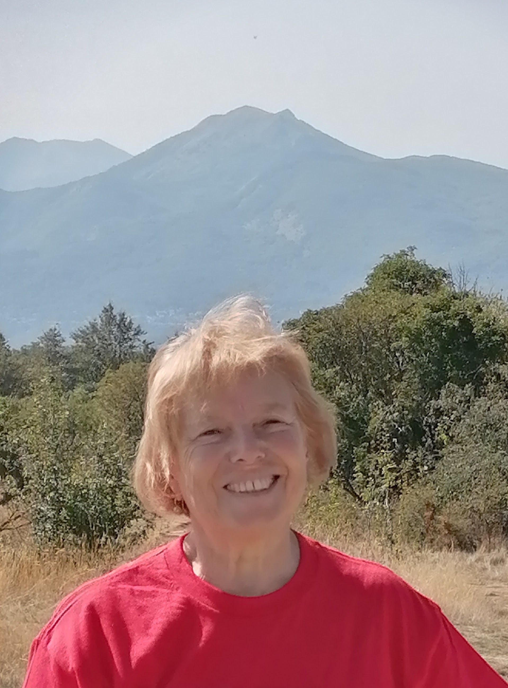 Person in a red shirt and blond hair smiling outside with trees and mountains in the background.