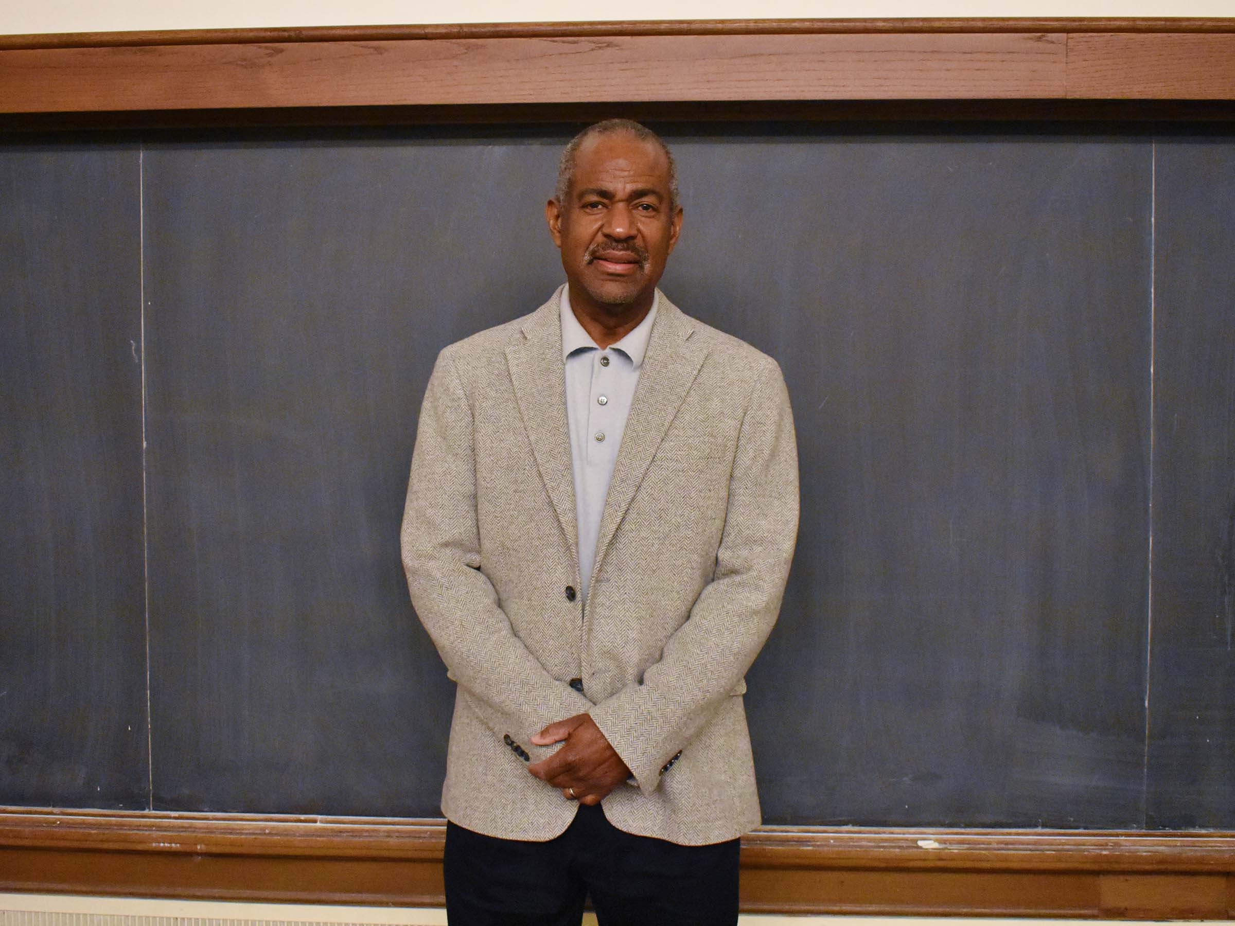 Dr. Michael Gomez, wearing a light colored, collared shirt, beige jacket and black pants, standing in front of a blackboard.