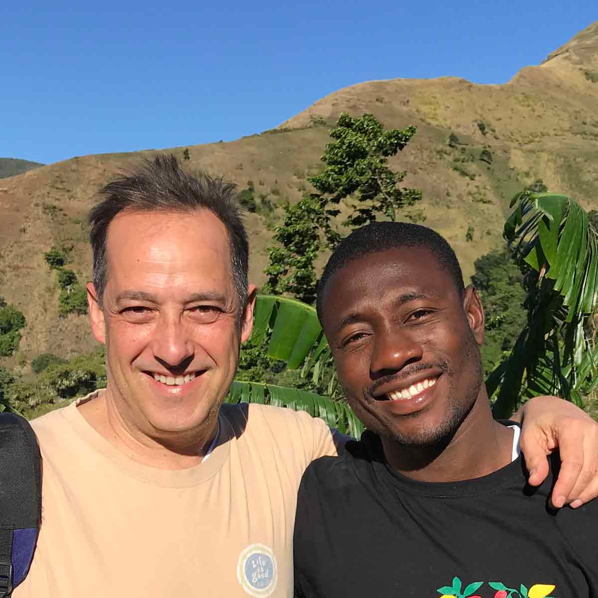 Two smiling people arm in arm in front of a mountain ridge.