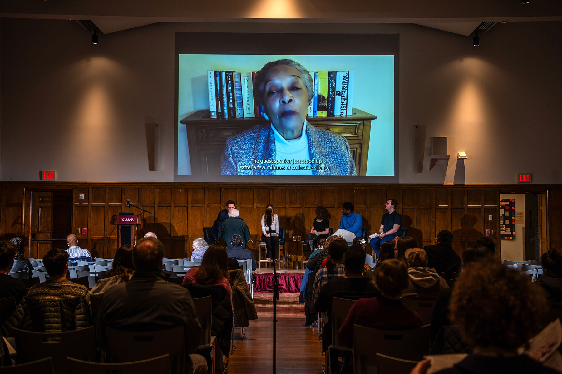 A panel of five people sitting in chairs on a stage with a large audience sitting in chairs. All are looking at a projection screen with a person speaking in front of a computer from Zoom.