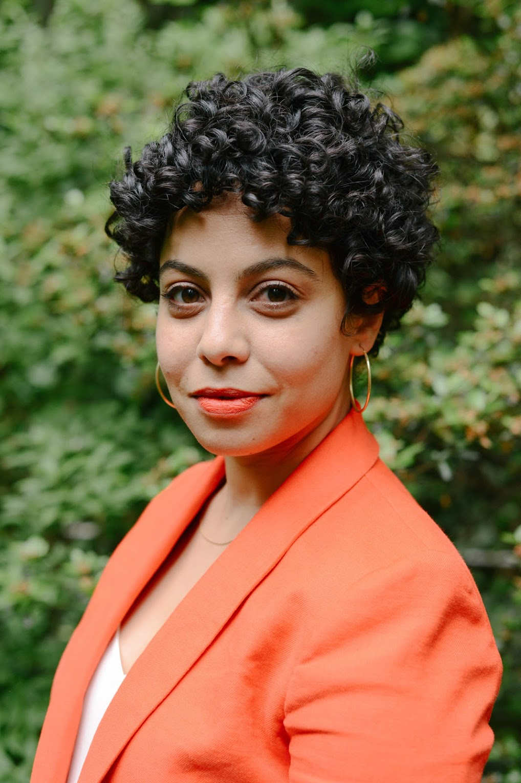 A person with short, black, curly hair and a bright orange suit jacket smiles at the viewer.