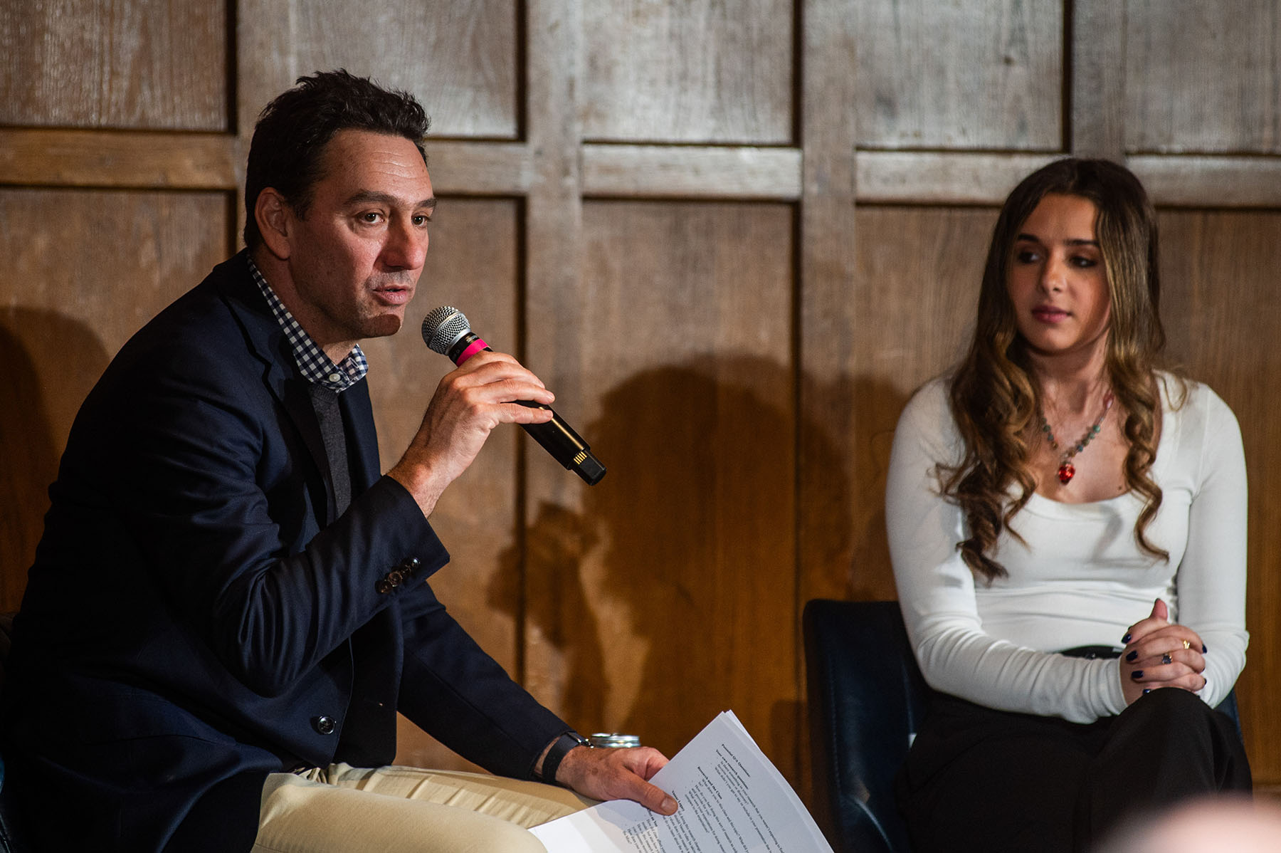 Two people sitting on a stage with a wood panel background. One person is speaking into a microphone while the other looks on.