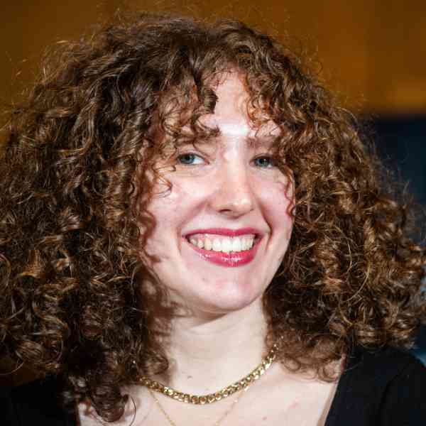 A person with long light-brown curly hair and a black shirt smiles at the camera.