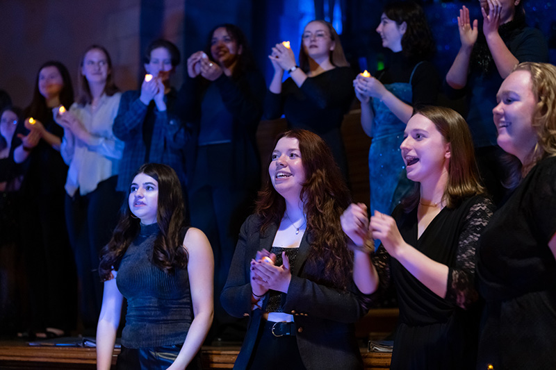 Performance of choir members standing, foreground row singing, background is a row of people holding a single candle each.