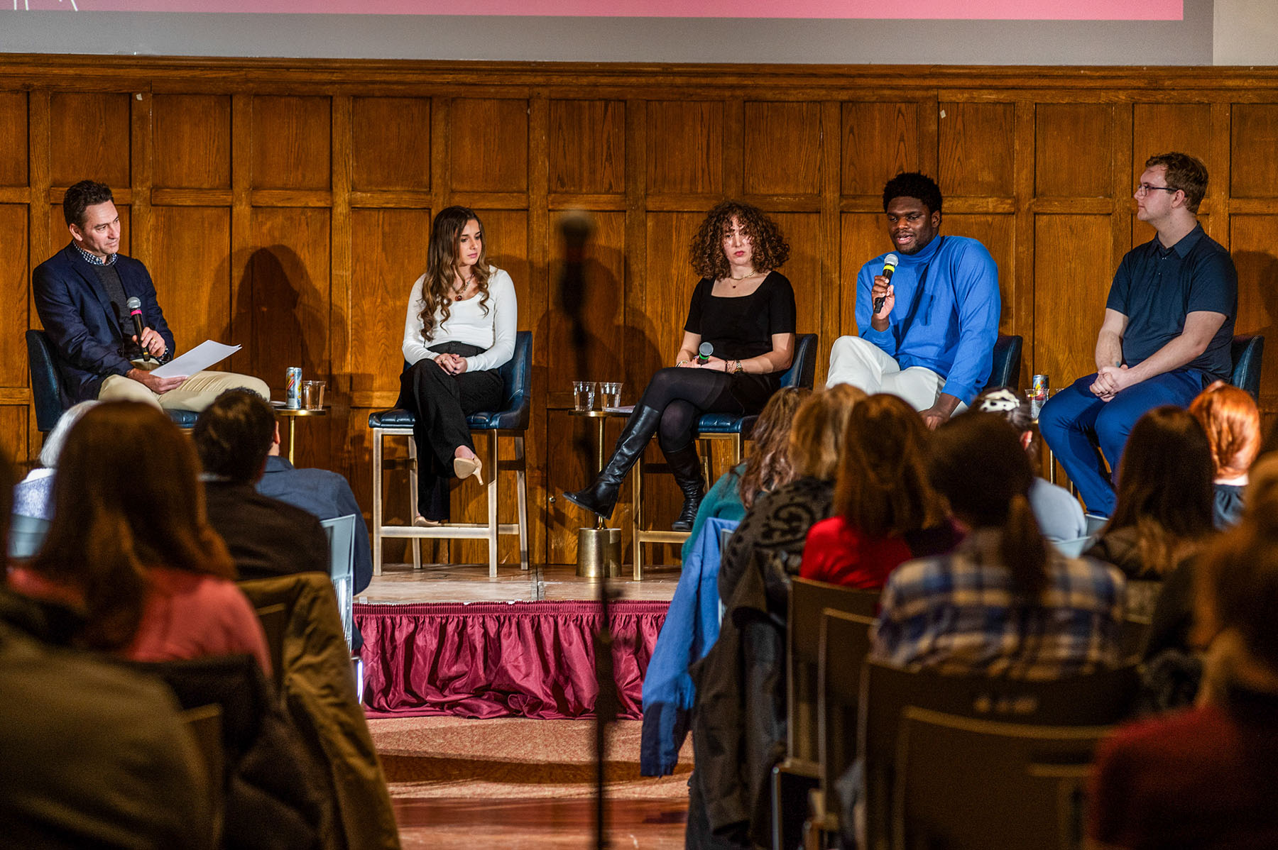 A panel of five people sitting in chairs on a stage with microphones speaking in front of an audience. 