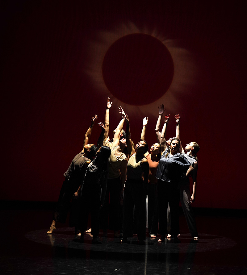 Nine dancers grouped with arms raised in a pose as if they were reaching for and facing the sun.