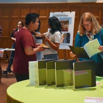 Two people looking at literature on a table in a large, wood-paneled room with other people in it.