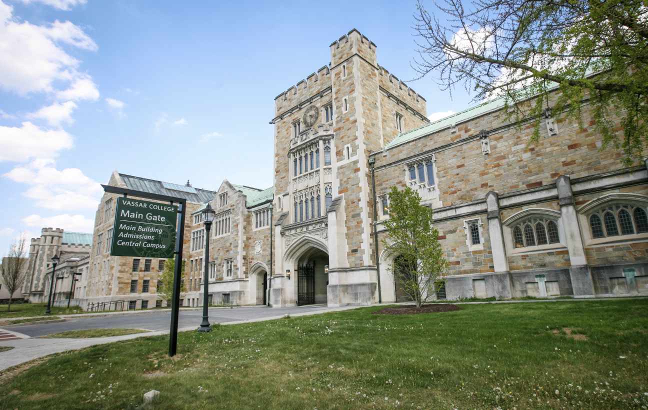 A photo of a large stone building with domed glass windows and a large open gate that allows cars to drive under the building. There is a grassy area in front of the building. A sign in front of the building reads “Vassar College Main Gate: Main Building, Admissions, Central Campus”.