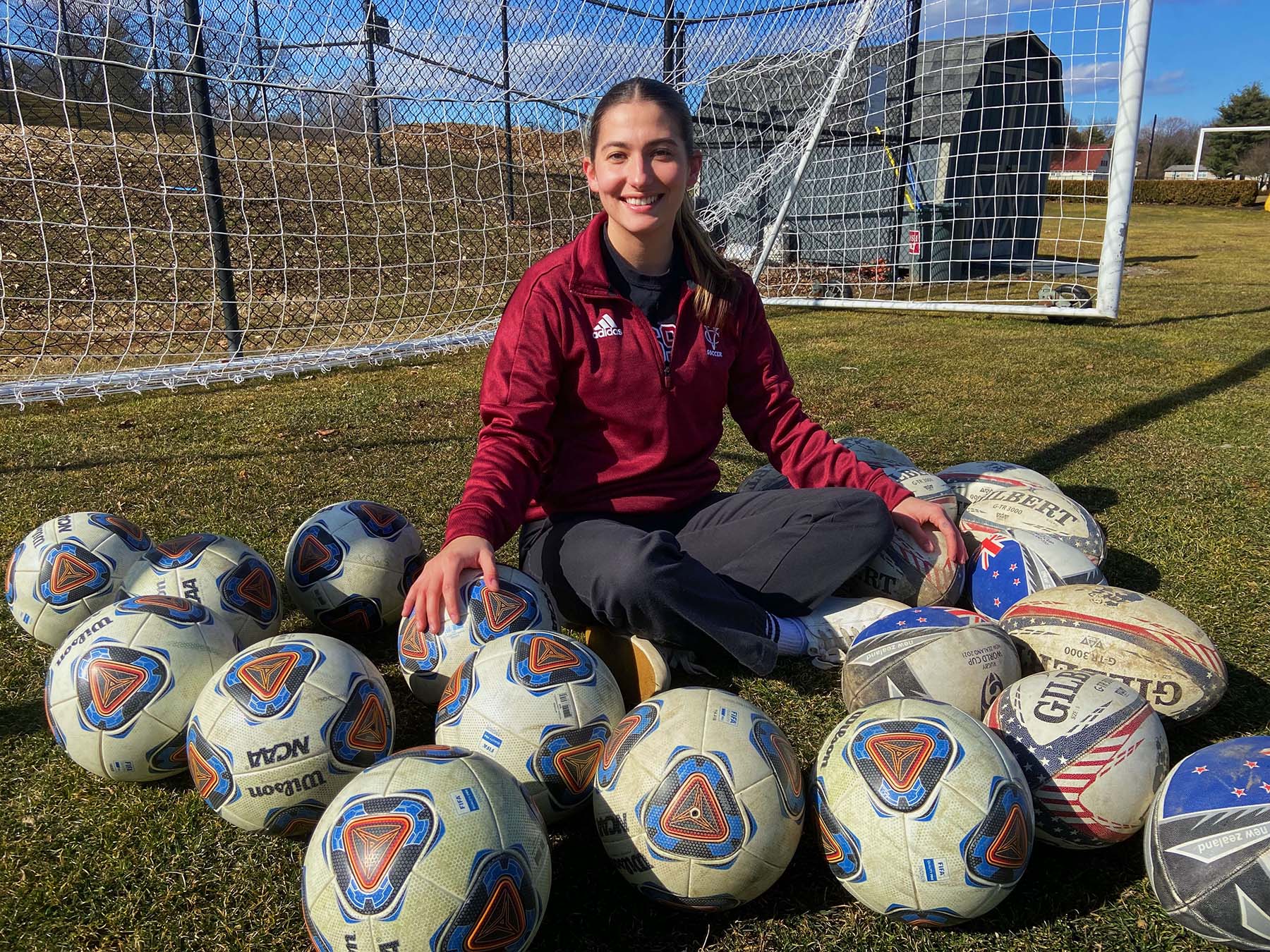 Person sitting on a soccer field in front of the goal surrounded my a bunch of soccer balls.