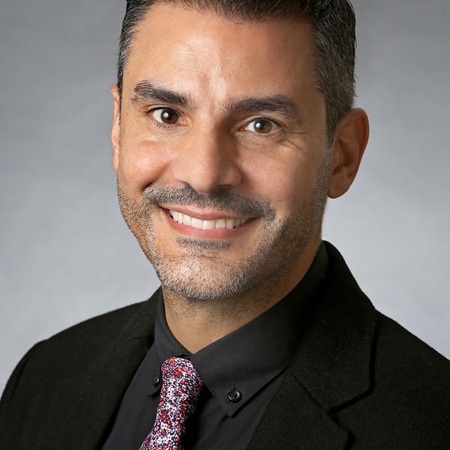 A portrait of a person with short black hair, facial hair, and black clothing and a magenta tie, smiling at the viewer.